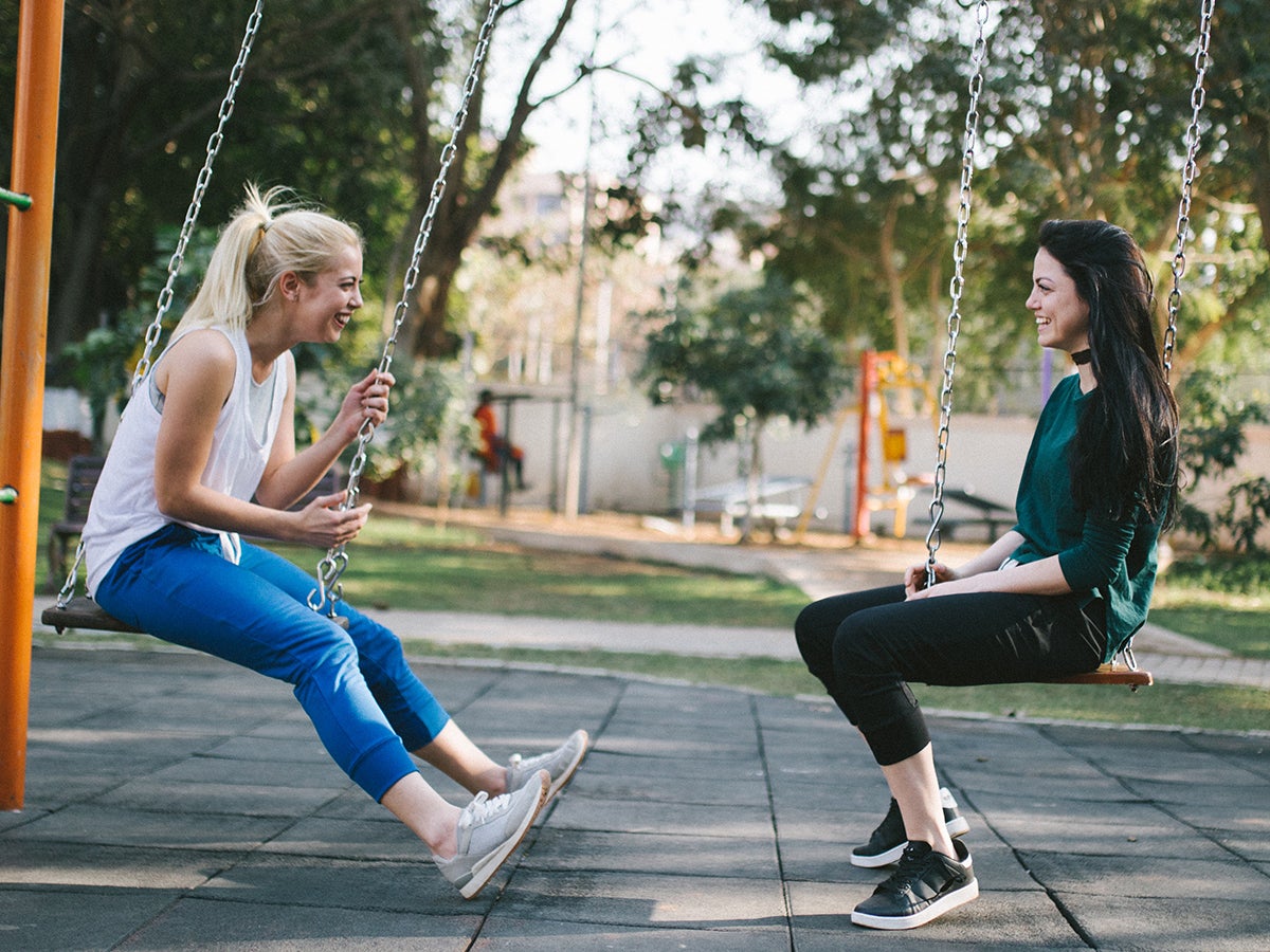 Dos mujeres sentadas cara a cara en un columpio en un parque infantil hablando entre sí. Una es rubia y usa jeans y una camisa blanca, la otra es morena y usa pantalones negros y una camisa verde.